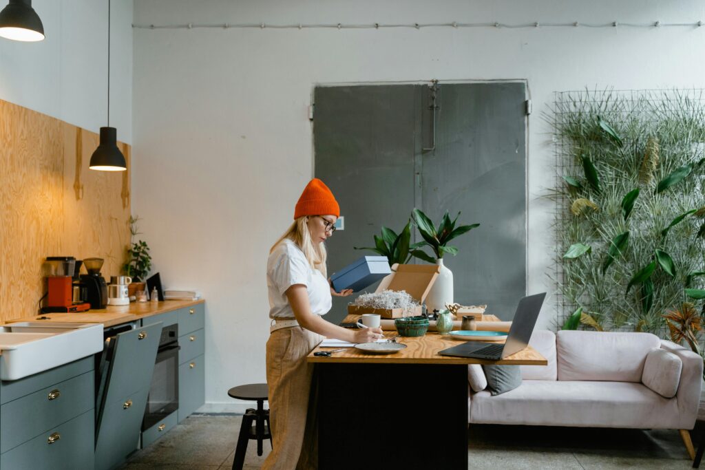 A young woman wearing an orange hat packaging products at her home office space with a laptop and green decor.