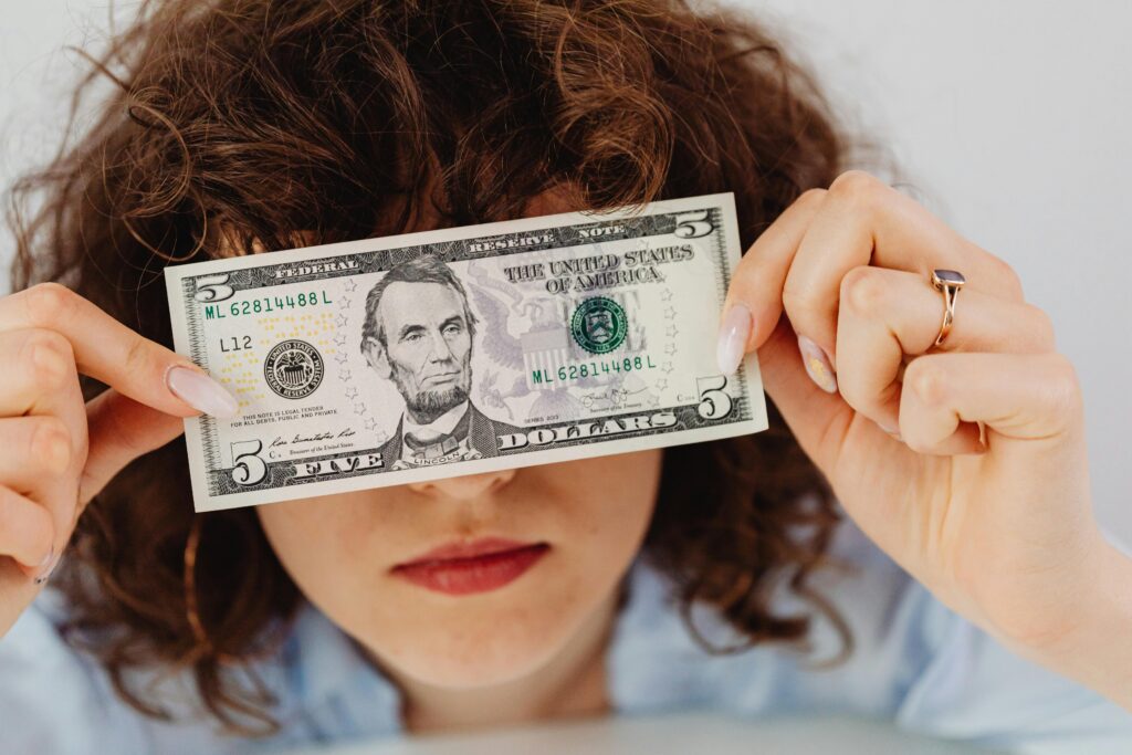 Woman with curly hair playfully covering eyes with a five dollar bill.